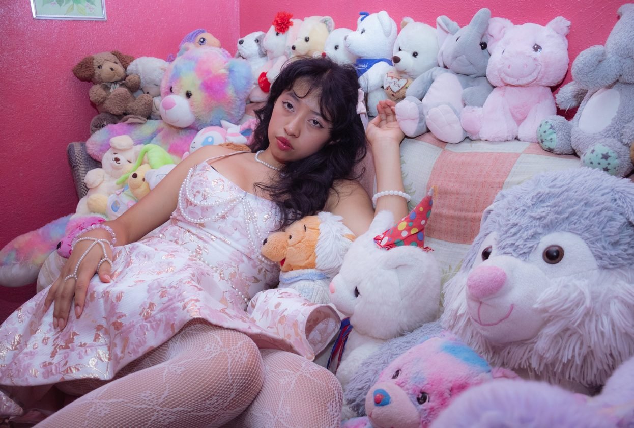 Retrato de una joven con vestido floral rodeada de peluches sobre una pared rosa